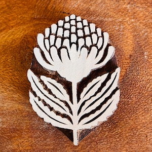 Puede incluir: Un bloque de madera tallada con un diseño floral. El diseño presenta una flor grande con pétalos y hojas detallados. El bloque es de madera marrón oscuro y la flor es de color crema claro. El bloque probablemente se utiliza para imprimir o estampar.