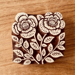 May include: A wooden block printing stamp featuring a floral design. The stamp depicts two white flowers with intricate details and leaves on a brown background.