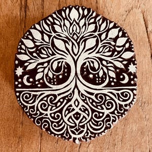 May include: A circular wooden printing block featuring a deeply carved design of a tree with intricate branches and roots. The raised surface is white, contrasting against the dark brown recessed areas, showcasing celestial motifs like stars and a crescent moon.