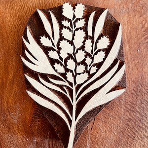 Puede incluir: Un bloque de madera con un diseño botánico. El bloque presenta un arreglo floral estilizado con hojas y un grupo central de pequeñas flores. El diseño es blanco sobre un fondo marrón oscuro. El bloque está sobre una superficie de madera.