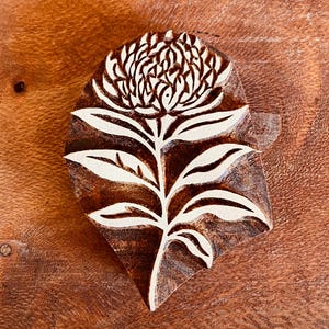 Puede incluir: Un bloque de madera con un diseño floral. El diseño presenta una flor y hojas detalladas, con detalles blancos sobre la madera marrón. Probablemente utilizado para imprimir o estampar.