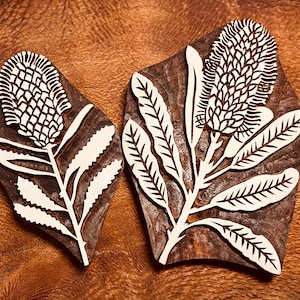 Op de afbeelding: Twee houten Banksia bloem drukvormen. De blokken zijn donkerbruin met een wit bloemmotief. De Banksia bloem en bladeren zijn in het hout gesneden. De blokken liggen op een houten oppervlak.