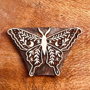 May include: A wooden butterfly stamp with intricate white details. The butterfly design features floral and celestial elements, including leaves, stars, and crescent moons. The stamp is set against a brown, textured background, showcasing the detailed carving.