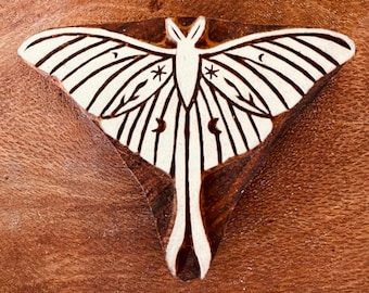Può includere: Un timbro in legno con un disegno di falena Luna. La falena ha ali bianche con strisce nere, stelle e dettagli a mezzaluna. Il timbro è posto su una superficie di legno marrone.