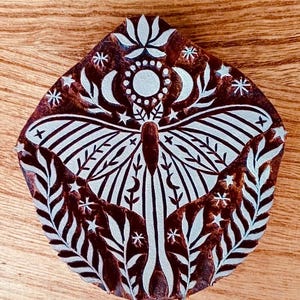 Puede incluir: Un sello de impresión de madera con un diseño blanco de una polilla con las alas extendidas, rodeado de estrellas, lunas y elementos florales. El fondo del sello es marrón.