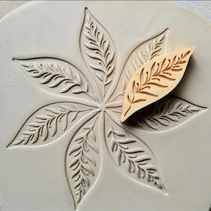 May include: A light gray, circular mold with a leaf pattern and a small, wooden leaf-shaped stamp. The mold features a detailed, raised design of a large, stylized leaf with intricate veins. The wooden stamp has a matching leaf design.