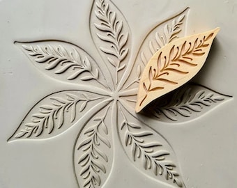 May include: A light grey, circular mould featuring a leaf pattern, alongside a small, wooden leaf-shaped stamp. The mould has a detailed, raised design of a large, stylised leaf with intricate veins. The wooden stamp has a matching leaf design.