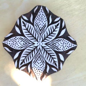Puede incluir: Un bloque de madera marrón oscuro con un diseño floral blanco. El diseño presenta una flor central con pétalos detallados y acentos punteados. El bloque probablemente se utiliza para imprimir o estampar.