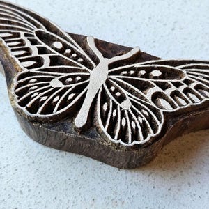 May include: A dark brown wooden block stamp featuring a carved butterfly design. The raised surface of the butterfly is light in color, showcasing intricate wing patterns with geometric shapes and lines carved into the wood grain texture.