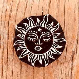 Peut inclure: Un tampon circulaire brun foncé avec un motif de visage de soleil blanc. Le soleil a les yeux fermés, une petite étoile et une bouche souriante. Les rayons du soleil sont stylisés avec des bords pointus. Le tampon est sur une surface en bois.