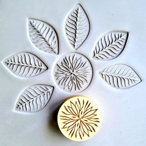 May include: A floral design stamp and impressions. The stamp is a light tan colour with a detailed flower design. The impressions are of leaves and a flower, all in white, arranged in a circular pattern on a white surface.