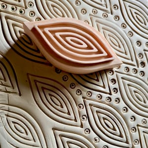 Puede incluir: Un sello de arcilla en forma de hoja, de color marrón claro, con patrones ovales concéntricos, que descansa sobre una superficie de arcilla de color crema con un diseño repetido de hojas y puntos. El sello probablemente se utiliza para crear patrones texturizados.