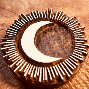 May include: A round, wooden decorative piece featuring a crescent moon design in the center. The moon is surrounded by a ring of vertical, white-painted lines, resembling sun rays. The piece is made of dark brown wood.