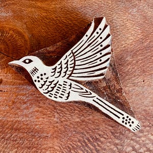 Könnte beinhalten: Ein dreieckiger Holzdruckblock mit einem geschnitzten Vogel im Flug. Das Vogelmotiv ist erhaben und hell gefärbt und zeigt detaillierte Flügelfedern, einen spitzen Schnabel und einen langen Schwanz mit kreisförmigen Mustern auf einem dunklen Holz.