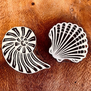 May include: Two wooden printing blocks featuring carved designs of a spiral nautilus shell and a fan-shaped scallop shell. The raised surfaces of the stamps are dark brown, while the recessed areas are light, creating a high-contrast graphic pattern.