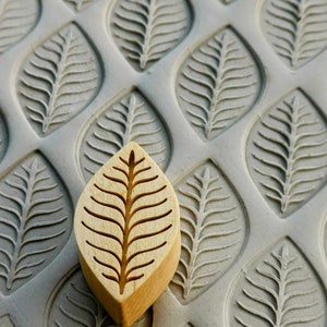 Puede incluir: Un sello de madera con forma de hoja con un diseño detallado de vetas de hoja se muestra sobre una superficie gris con un patrón de hojas. El sello es de color marrón claro y el patrón de la superficie es de color gris claro. La imagen destaca el intrincado diseño del sello.