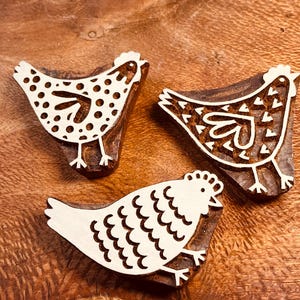 Puede incluir: Tres sellos de madera con forma de gallina con diseños blancos sobre un fondo marrón. Cada sello presenta un diseño de gallina diferente, con detalles punteados, estampados y festoneados. Ideal para manualidades y un toque rústico.