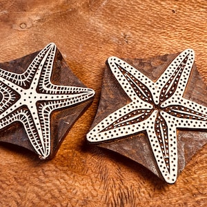 May include: Two wooden starfish stamps with intricate white designs, resting on dark, square bases. The starfish have detailed patterns and are set against a warm, wooden background, showcasing a craft or art supply.