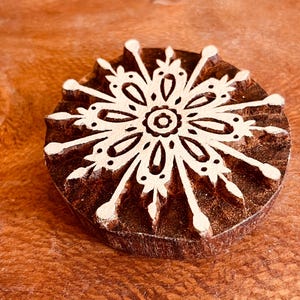 May include: A round, wooden block with a detailed snowflake design carved into its surface. The snowflake is white against the dark brown wood, showcasing intricate patterns and shapes. This craft supply is ideal for printing or stamping.