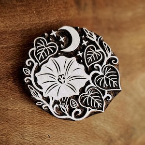 May include: A circular wooden printing block featuring a carved design of a large morning glory flower surrounded by heart-shaped leaves, a crescent moon, and small stars. The raised relief pattern is white against the dark brown wood surface.