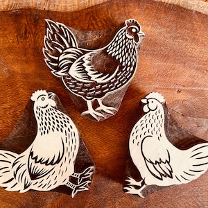 Puede incluir: Tres bloques de impresión de madera tallada, cada uno con un diseño de pollo distinto. Los bloques están hechos de madera marrón oscura con superficies elevadas de color claro que forman los contornos y detalles de las plumas.