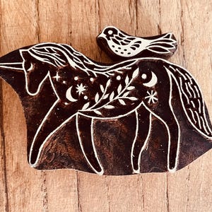 May include: A dark brown wooden block featuring a unicorn design with white accents. The unicorn has a flowing mane, and the design includes stars, moons, and floral elements. A small bird sits on the unicorn's back.
