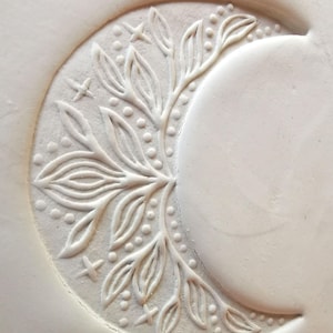 May include: A white, crescent moon design with embossed floral and star details. The design features a central branch with leaves and dotted accents, along with small star shapes. The moon's surface is smooth, contrasting with the textured details.