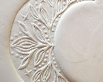 May include: A white, crescent moon-shaped design with embossed floral and star details. The design features a central branch with leaves and dotted accents, along with small star shapes. The moon's surface is smooth, contrasting with the textured details.