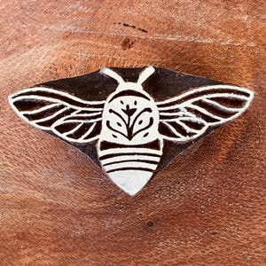May include: A wooden stamp featuring a bee design. The bee has white wings and body details on a black background. The stamp is set on a brown wooden surface.