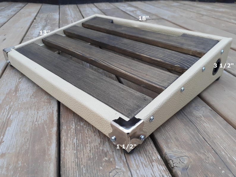 K&ouml;nnte beinhalten: Ein rechteckiges Pedalboard mit cremefarbenem Rahmen und dunklen Holzlatten. Das Pedalboard ist 45,7 cm lang, 33 cm breit und 8,9 cm hoch. Die Ecken sind mit silberfarbenem Metall verst&auml;rkt. Der Rahmen ist 3,8 cm dick.