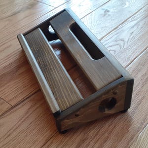 May include: A rectangular wooden pedalboard with a dark brown stained finish. The structure features two horizontal slats with a central gap and side panels containing circular cutouts. The design includes a raised front edge and a slanted profile.