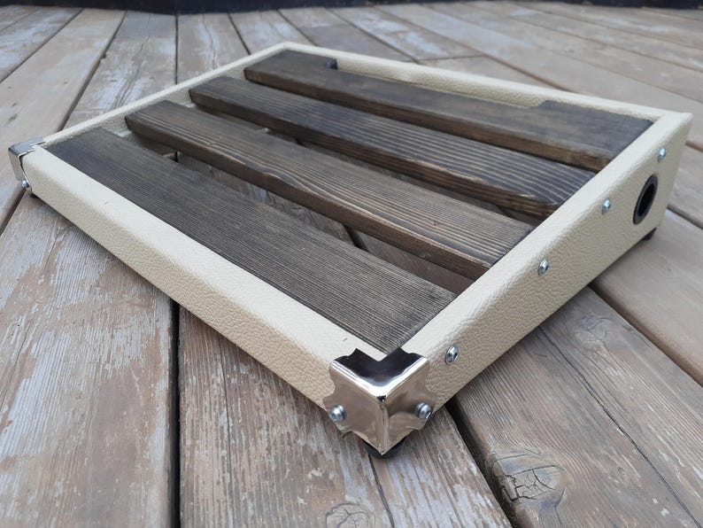 K&ouml;nnte beinhalten: Ein beigefarbenes und braunes Pedalboard aus Holz mit rechteckiger Form. Es hat drei dunkelbraune Holzlatten und einen beigefarbenen Rahmen mit silbernen Ecksch&uuml;tzern. Auf der Seite ist eine schwarze Eingangsbuchse zu sehen.