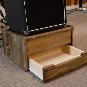 May include: A rectangular wooden stand with a dark brown stained finish and a single pull-out drawer featuring a light-colored interior. The stand has visible metal fasteners on the side panels and supports a black amplifier on its top surface.