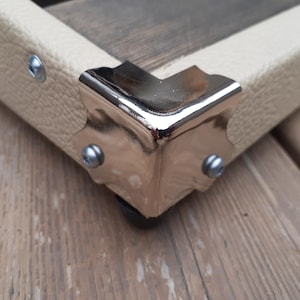 K&ouml;nnte beinhalten: Nahaufnahme einer Ecke einer Holzkiste mit cremefarbenem Kunstlederbesatz. Die Ecke ist mit einem gl&auml;nzenden, silberfarbenen Metalleckenschutz verst&auml;rkt, der mit kleinen silbernen Nieten befestigt ist. Die Kiste ruht auf einer Holzoberfl&auml;che.