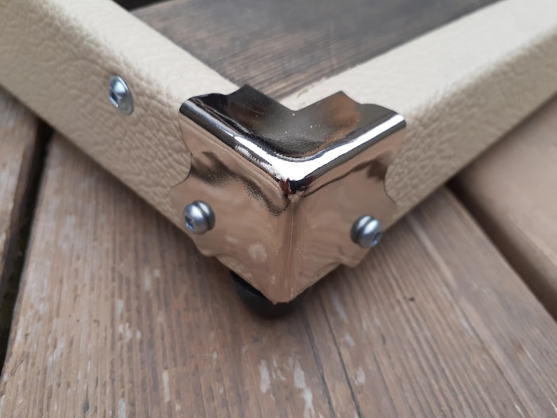 K&ouml;nnte beinhalten: Nahaufnahme einer Ecke einer Holzkiste mit cremefarbenem Kunstlederbesatz. Die Ecke ist mit einem gl&auml;nzenden, silberfarbenen Metalleckenschutz verst&auml;rkt, der mit kleinen silbernen Nieten befestigt ist. Die Kiste ruht auf einer Holzoberfl&auml;che.