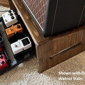 May include: A dark walnut stained wooden amp stand with a built-in pedalboard. The pedalboard holds several guitar effects pedals in various colors. The amp stand supports a black amplifier with a brown speaker grill. Text on the image reads "Shown with Dark Walnut Stain."