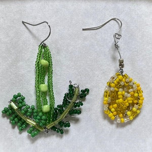 Puede incluir: Un par de pendientes de cuentas. Un pendiente es verde y amarillo y el otro es amarillo. Los pendientes están hechos de cuentas pequeñas y tienen forma de hojas.