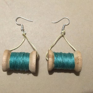 May include: A pair of earrings featuring wooden spools with teal thread wrapped around them. The spools are suspended from silver hooks with a light yellow thread.