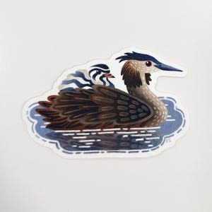 May include: A sticker featuring a stylized illustration of a Great Crested Grebe swimming in blue water. The bird has brown, black, and white feathers, a blue beak, and a red eye. A smaller bird is riding on its back.
