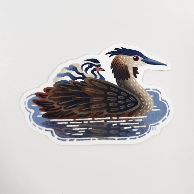 May include: A sticker featuring a stylized illustration of a Great Crested Grebe swimming in blue water. The bird has brown, black, and white feathers, a blue beak, and a red eye. A smaller bird is riding on its back.