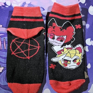 May include: A pair of black ankle socks with red trim. One sock features a red pentagram and the other features a cartoon image of two characters with red and white accents.