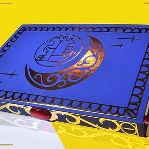 May include: A blue hardcover book with a gold decorative border and a gold crescent moon design with a symbol inside. The book has a red jewel on the spine and a purple ribbon bookmark.