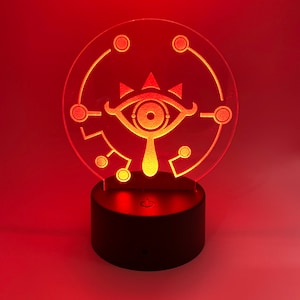 May include: A red LED night light with a clear acrylic base featuring a white silhouette of the Triforce symbol from the Legend of Zelda video game series. The light is on a black base with a switch.