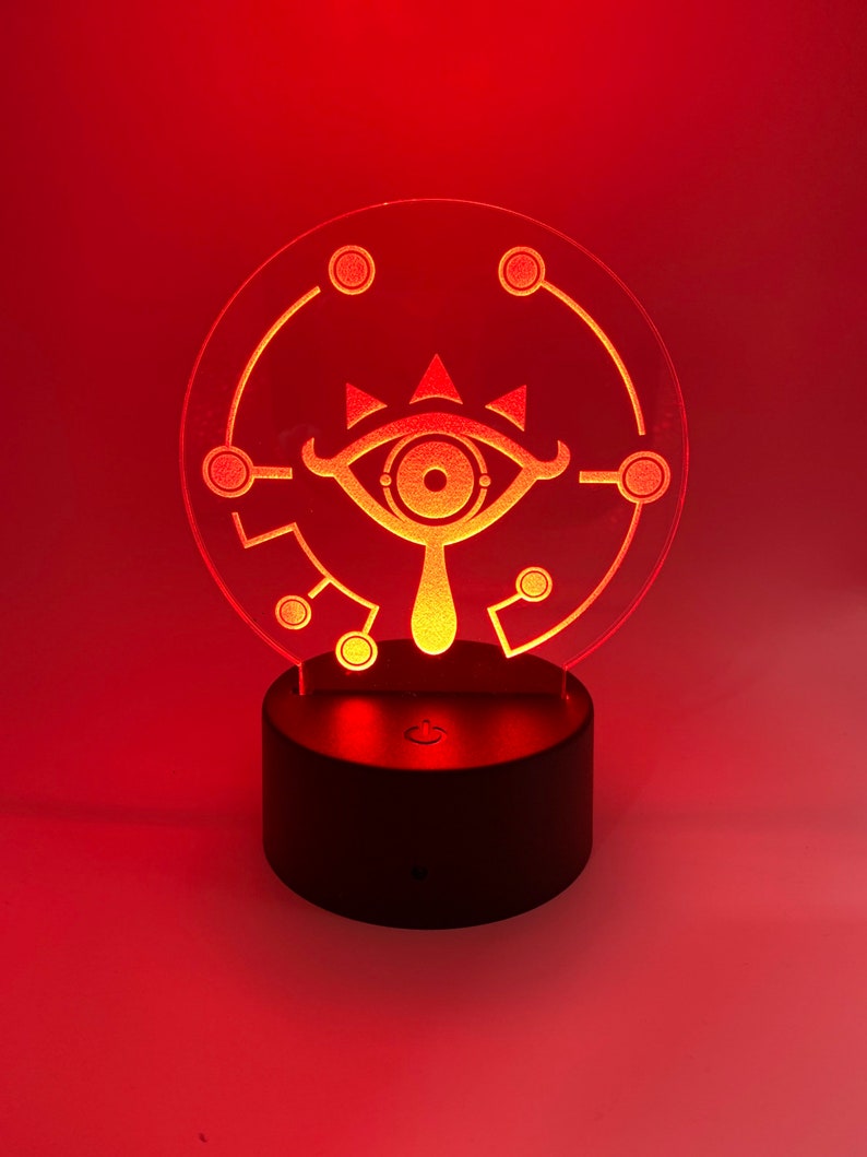 May include: A red LED night light with a clear acrylic base featuring a white silhouette of the Triforce symbol from the Legend of Zelda video game series. The light is on a black base with a switch.