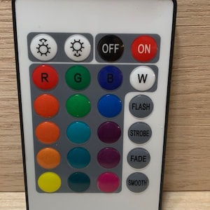 May include: A white remote control with black buttons and a black border. The buttons are arranged in a grid with the top row having buttons labeled "OFF" and "ON". The second row has buttons labeled "R", "G", "B", and "W". The remaining rows have buttons labeled "FLASH", "STROBE", "FADE", and "SMOOTH".