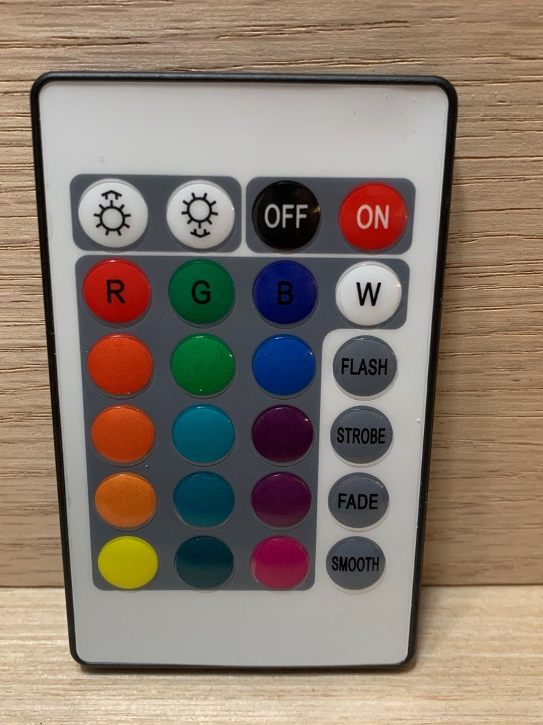 May include: A white remote control with black buttons and a black border. The buttons are arranged in a grid with the top row having buttons labeled "OFF" and "ON". The second row has buttons labeled "R", "G", "B", and "W". The remaining rows have buttons labeled "FLASH", "STROBE", "FADE", and "SMOOTH".