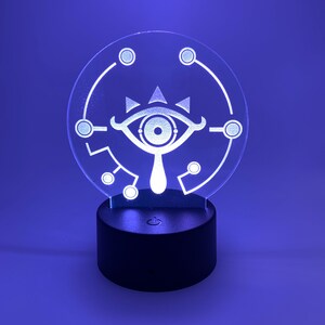 May include: A white 3D printed lamp with a circular base and a clear acrylic panel featuring a stylized eye design. The lamp is lit up with a blue light.
