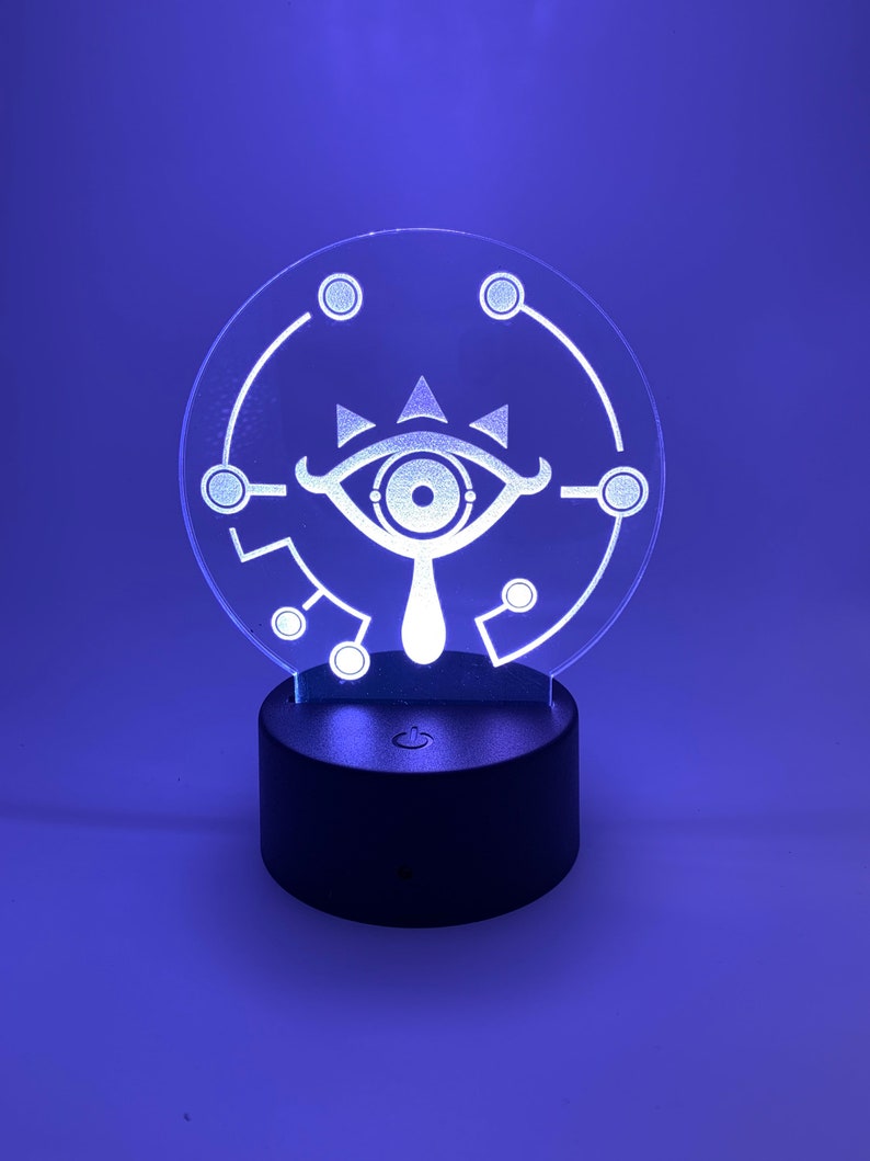 May include: A white 3D printed lamp with a circular base and a clear acrylic panel featuring a stylized eye design. The lamp is lit up with a blue light.