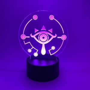 May include: A white 3D printed night light shaped like the Sheikah Eye from the Legend of Zelda video game series. The light is on a black base and is glowing purple.