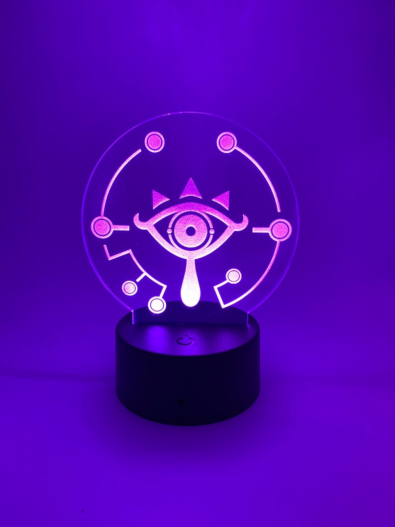 May include: A white 3D printed night light shaped like the Sheikah Eye from the Legend of Zelda video game series. The light is on a black base and is glowing purple.
